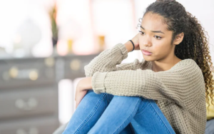girl in olive top and blue jeans seated on ground looking sad